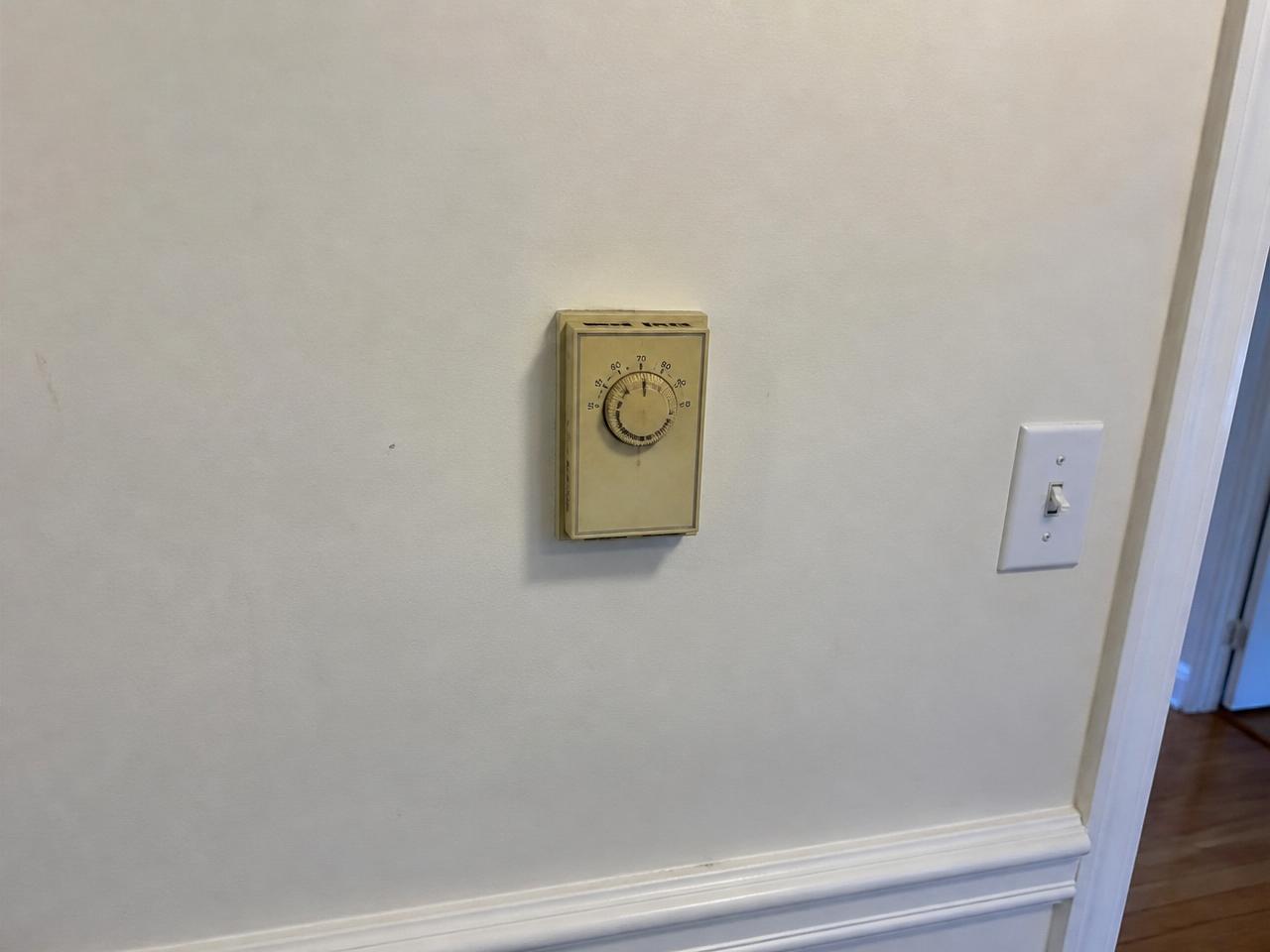Before: Same-location before: old yellowed manual dial thermostat on an interior hallway wall